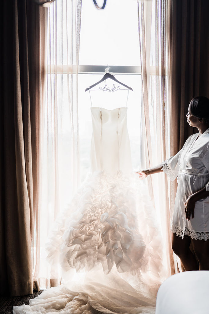 Wedding dress on hanger with backlighting and bride admiring it 
