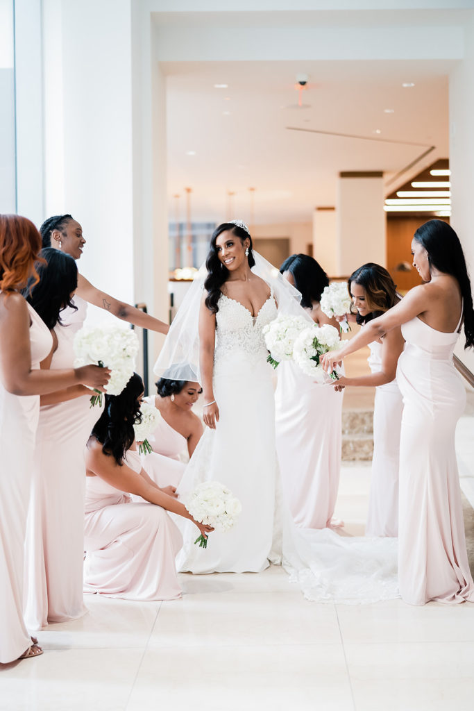Bridal party getting ready photo with the bride admiring her dress in solid light pink floor length gowns with ribbon sash