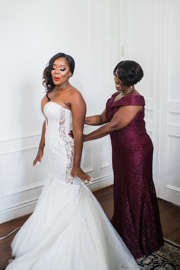 Mother of bride and bride putting on wedding dress while getting ready. Mother of bride in burgundy fitted floor length dress and bride in strapless wedding dress with plunging neckline and lace detailing 